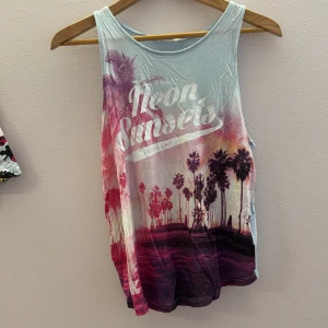 Linnetopp med tryck 'Neon Sunsets' - Säljer en cool linnetopp med trycket 'Neon Sunsets Every Day'. Toppen har ett färgglatt motiv med palmer i rosa och lila toner. Perfekt för sommarens varma dagar. är i ungdom storlek 170 14+