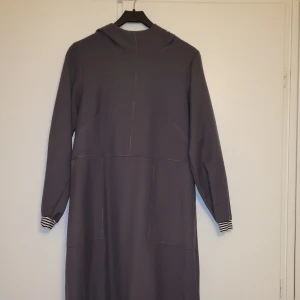 Grå långärmad klänning med huva - Snygg grå klänning med huva och långa ärmar. Den har en enkel design med randiga detaljer vid ärmsluten. Perfekt för en avslappnad stil. Sydd i ett vävt ylletyg. 