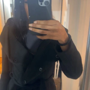 Svart kappa med knappar - Snygg kort, svart kappa med knappar, bälte i midjan och huva . Perfekt för kyligare dagar med sin stilrena design och långa ärmar.  Använt några gånger 