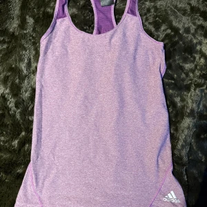 Lila träningslinne från Adidas - Säljer ett snyggt lila träningslinne från Adidas. Linnet har en brottarrygg och är tillverkat i ett mjukt och stretchigt material som andas. Perfekt för gymmet eller löprundan!