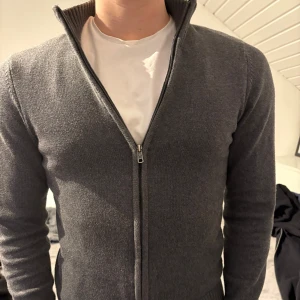 Grå kofta från J.F.K - Säljer en stilren grå kofta från J.F.K i 100% pima bomull. Koftan har en dragkedja framtill och är ribbad vid krage, ärmslut och nederkant. Perfekt för en avslappnad men ändå snygg look.