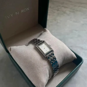 Silverfärgad klocka från Ivy N Rox - silverfärgad klocka från Ivy N Rox med rektangulär urtavla.  Klockan har en länkarmband i metall och stenar runt om urtavlan. Jag har tagit av två länkar men dem skickas med!  💕