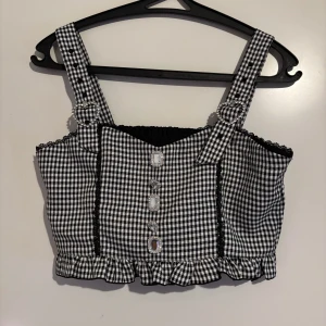 Svartvit rutig korsettopp - Säljer en snygg svartvit rutig korsettopp från Dear My Love. Toppen har breda axelband och dekorativa knappar framtill. Den är croppad med volangdetaljer vid nederkanten och har en elastisk rygg för bättre passform. Passar perfekt till jirai kei och ryousangata!