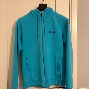 Turkos hoodie från Boss - Säljer en snygg turkos hoodie från Boss med dragkedja och huva. Tröjan har långa ärmar och praktiska fickor framtill. Perfekt för en avslappnad stil.
