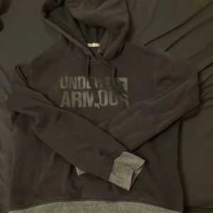 Svart hoodie från Under Armour - Säljer en svart hoodie från Under Armour med grått tryck på framsidan. Tröjan har en klassisk design med huva och långa ärmar. Perfekt för en avslappnad stil.