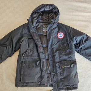 Svart dunjacka från Canada Goose - Säljer en svart dunjacka från Canada Goose med huva och två stora fickor framtill. Jackan har en broderad logotyp på ärmen och är perfekt för kalla vinterdagar. Den läcker lite fjädrar dock!