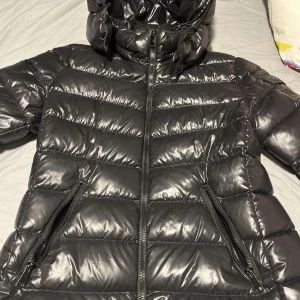 Svart dunjacka från Moncler - Snygg svart dunjacka från Moncler med glansig finish och huva. Jackan har dragkedja framtill och två fickor med dragkedjor. Perfekt för kyliga dagar när du vill hålla stilen. 🖤