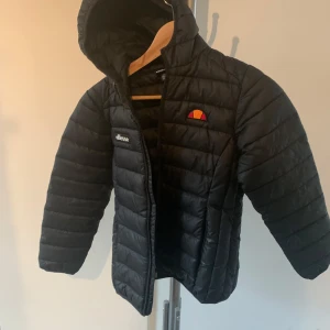 Svart dunjacka från Ellesse - Säljer en svart dunjacka från Ellesse med huva och dragkedja. Jackan har ett quiltat mönster och Ellesse-loggan på bröstet. Perfekt för kyliga dagar! Obs, dragkedja på fickorna är trasiga 