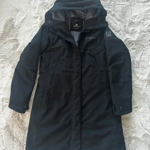 Svart parkajacka från Didriksons - Säljer en svart längre parkajacka från Didriksons med avtagbar fuskpäls på huvan. Jackan är 100% vattentät och har dragkedja framtill. Perfekt för kalla och blöta dagar. Fodret är gjort av polyester för extra värme.