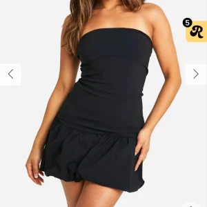 Svart axelbandslös klänning - Snygg svart axelbandslös klänning med en kort och figurnära passform. Perfekt för en kväll ute eller en speciell tillställning. Den har en volangdetalj vid fållen som ger en extra touch av stil.