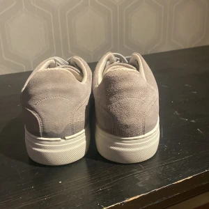 Grå sneakers från Selected Homme - Snygga grå sneakers i mocka från Selected Homme. De har en stilren design med snörning och en vit sula som ger en fräsch look. Perfekta för en avslappnad stil.