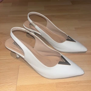 Vita slingback pumps med spetsig tå - Eleganta vita slingback pumps med spetsig tå och dekorativ metalltriangel framtill. Perfekta för en stilren look. Klackhöjden ger en bekväm passform.