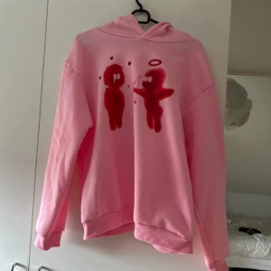 Rosa hoodie med tryck - Säljer en rosa hoodie med ett unikt tryck av två figurer på framsidan. Hoodien har en klassisk passform med långa ärmar och en bekväm huva. Perfekt för en avslappnad stil.