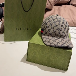 Gucci keps blå - HELT NY. Snygg beige keps från Gucci med det ikoniska GG-mönstret. Kepsen har en justerbar rem baktill och en röd och blå randdetalj på sidan. Perfekt för att ge din outfit en lyxig touch.