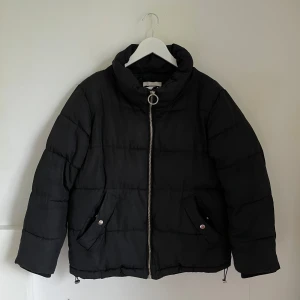 Svart pufferjacka från H&M - Säljer en svart pufferjacka från H&M i storlek S. Jackan har en cool dragkedja med en stor ringdetalj och två fickor framtill. Perfekt för kalla dagar när du vill hålla dig varm och snygg. Jackan är i bra skick och redo för en ny ägare!