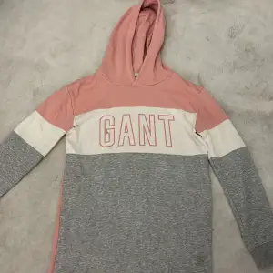 Jag säljer en superfin hoodie från GANT i rosa och grått med vit detalj. Den har en stor GANT-logga på framsidan och 'GIRLS' på baksidan. Perfekt för en avslappnad stil och mysiga dagar. Hoodien har långa ärmar och är gjord i mjukt material.