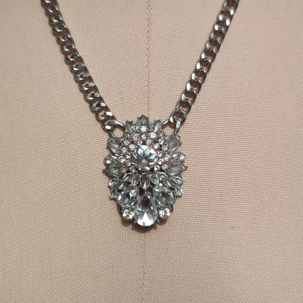 Säljer ett snyggt silverfärgat halsband med en stor glittrig detalj i mitten. Kedjan är robust och ger en cool look, perfekt för att piffa upp vilken outfit som helst. Detaljen i mitten är formad som en blomma med flera gnistrande stenar. Passar perfekt till fest eller en utekväll!. Accessoarer.