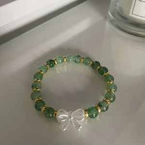 Grönt armband med rosett - Snyggt armband med gröna pärlor och gulddetaljer. Armbandet har en söt rosett i transparent material som ger en extra touch. Perfekt för att piffa upp vilken outfit som helst!