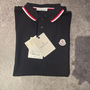 Svart moncler piké  - Säljer en svart Monclear piké helt ny och oanvänd med etikett, qr kod och påse. Vid minsta fundering är det bara att höra av sig! Nypris 3000kr men säljer den för  1500kr.
