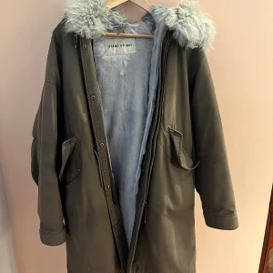 Olivgrön parkajacka med päls  från Stand Studio - Snygg olivgrön parkajacka från Stand Studio med mjukt foder och päls i cashmere . Jackan har en dragkedja och knappar framtill samt praktiska fickor. Perfekt för kyliga dagar. Går att ta bort ytlager för tvätt