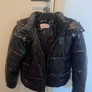 Svart pufferjacka från Nelly - Snygg svart pufferjacka från Nelly med glansig finish. Jackan har en dragkedja framtill och en praktisk huva. Perfekt för kyligare dagar med sin vadderade design.