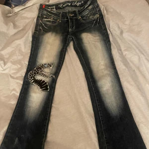 Mörkblå jeans med ormmönster från Crazy Age - Snygga mörkblå jeans från Crazy Age med ett coolt ormmönster på ena benet. De har en låg midja och en bootcut-stil. storlek 36/26 och de sitter perfekt på mig som är drygt 160cm! dem är lite sönder i dragkedjan men det har jag fixat med ett gummiband. pris kan diskuteras!
