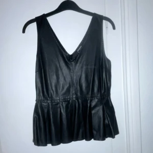 Svart skinntopp med v-ringning - Snygg svart skinntopp med v-ringad design och volangdetaljer i nederkant. Perfekt för att ge en edgy touch till din outfit. Jättefint skick✨
