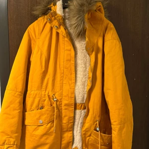 Gul parkajacka med fuskpäls - Snygg gul parkajacka med fuskpäls på huvan. Jackan har dragkedja och knappar framtill samt praktiska fickor med knappar. Perfekt för kyliga dagar med sin varma och mysiga insida.
