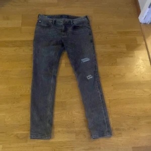 Grå jeans från Scotch & Soda - Snygga grå jeans från Scotch & Soda med slitna detaljer på framsidan. De har en klassisk femficksdesign och en läderpatch med logotyp på baksidan. Perfekta för en avslappnad stil.