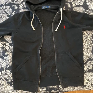 Svart hoodie från Polo Ralph Lauren - Säljer en svart hoodie från Polo Ralph Lauren med dragkedja och luva. Den har en röd logotyp på bröstet och vita snören vid huvan.   FAST PRIS