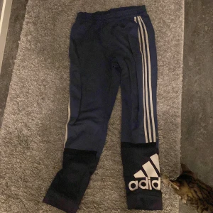 Träningsbyxor från Adidas - Säljer två par träningsbyxor från Adidas. Det första paret är blått med vita ränder och stor logga vid vristen. Det andra paret är svart med orange detaljer och snörning i midjan. Perfekta för träning eller en avslappnad dag. Svarta paret är knappt avnvänd. Dem blå har använts men är i bra skick.