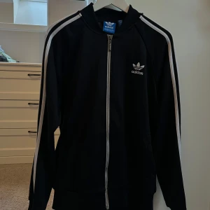 Svart träningsjacka från Adidas - Snygg svart träningsjacka från Adidas med vita ränder längs ärmarna och dragkedja framtill. Jackan har en klassisk design med Adidas-logga på bröstet och långa ärmar. Perfekt för sportiga aktiviteter eller en avslappnad stil.