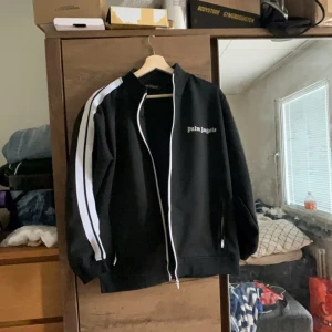 Svart track jacket från Palm Angels - Snygg svart track jacket från Palm Angels med vita ränder längs ärmarna. Jackan har dragkedja framtill och en stilren design med logotyp på bröstet. Perfekt för en sportig look.