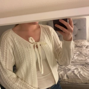 Vit stickad kofta  - Säljer en mysig vit stickad kofta med knytning framtill. Koftan har ett fint kabelstickat mönster och långa ärmar. Perfekt för kyliga dagar! Vet ej vart den är köpt. 