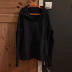 Mörkblå gant kofta med dragkedja - Säljer en stilren mörkblå gant kofta med dragkedja och hög krage. Koftan har långa ärmar och ribbade muddar vid ärmslut och nederkant. Perfekt för kyliga dagar!