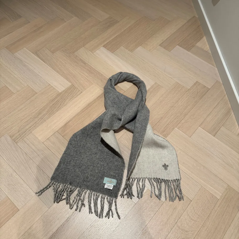 Snygg och stilren halsduk från Morris Lady i grått och vit. Tillverkad i 100% virgin ull och prydd med en liten broderad logotyp. Perfekt för att hålla dig varm under kyliga dagar. Tillverkad i Italien med fransar i ändarna för en klassisk look. Har tvättats en gång i handtvättsprogram och har blivit något krympt.. Asusteet.