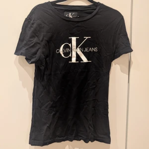 Svart t-shirt från Calvin Klein Jeans - Snygg svart t-shirt från Calvin Klein Jeans med det ikoniska CK-logotypen tryckt i vitt på framsidan. Perfekt för en stilren och avslappnad look. Finns lite slitningar på trycket, se bilder. Storlek XS. 