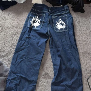 Blå jeans med yin-yang-motiv - Snygga blå jeans med unikt yin-yang-motiv och flammor på bakfickorna. Perfekta för dig som vill sticka ut med en cool och annorlunda stil. Jeansen har en klassisk passform och är tillverkade i ett slitstarkt material. (Storlek 40/42 - L) 