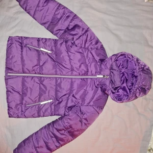 Lila pufferjacka vinter Ellos  - Säljer en snygg lila pufferjacka från Ellos i storlek 128. Jackan har en praktisk huva och dragkedja framtill. Perfekt för kyliga dagar med sin quiltade design och reflekterande detalj på baksidan. Förstärkt lägst ner vid dragkedjan och finns pyttesmå skavanker vid armen om man letar. Se bilder.         #barn #vinterjacka #tonåring 