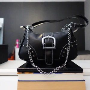 Svart axelväska med kedjedetaljer - Snygg svart axelväska i syntetmaterial med silverfärgade kedjedetaljer och en stor dekorativ spänne framtill. Väskan har en dragkedja upptill och kontrasterande vita sömmar som ger en cool look. Perfekt för att ge din outfit en edgy touch.