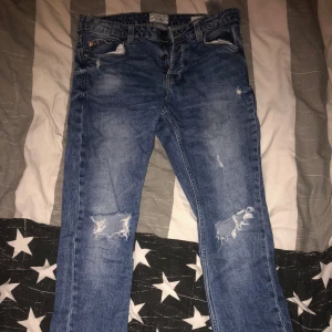 Blå jeans med slitningar, slim jeans - Säljer ett par blå jeans med coola slitningar på knäna. De har en klassisk femficksdesign och knappgylf. Perfekta för en avslappnad stil.