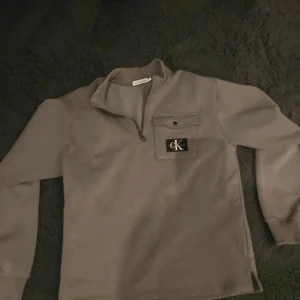 Grå tröja med dragkedja från Calvin Klein - Snygg grå tröja från Calvin Klein med dragkedja och en bröstficka med CK-logga. Perfekt för en stilren och avslappnad look. 
