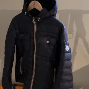 Svart dunjacka från Moncler - Snygg svart dunjacka från Moncler med huva och dragkedja. Jackan har två bröstfickor med knappar och sidofickor med dragkedja. Perfekt för kyliga dagar.