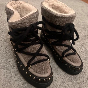 Boots med snörning från Inuikii - Säljer ett par snygga boots från Inuikii med svart snörning och nitar runt sulan. De är i ett ”fluffigt” material med skinnmaterial nertill. Strl 38. Tyvärr saknar de metallpiggarna i ändarna på snörena