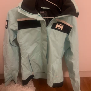 Ljusblå vindjacka från Helly Hansen - Snygg ljusblå vindjacka från Helly Hansen med dragkedja och hög krage. Jackan har svarta detaljer och logotyp på bröstet och ärmen. Perfekt för blåsiga dagar. Använd en gång