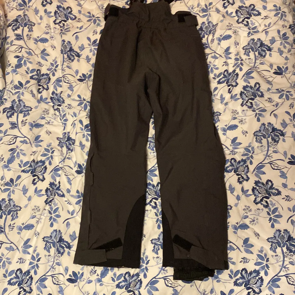 Säljer ett par svarta byxor från Bergans med justerbara hängslen. Byxorna är tillverkade i 100% polyester och har en praktisk design för utomhusaktiviteter. Perfekta för vinteräventyr med slitstarka detaljer och bra passform.. Farkut & Housut.