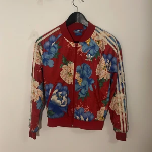Blommig röd jacka från Adidas Japan - Snygg röd Adidas ”track top” med stort blommönster. Jackan har dragkedja och de klassiska tre ränderna längs ärmarna. Perfekt för att sticka ut med en färgglad stil.  Liten storlek, ca XS, 150. 