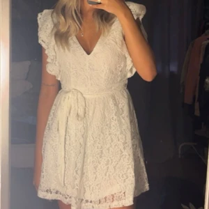 Vit spetsklänning med volanger - Säljer min vit studentklänning med volanger och öppen rygg. Klänningen har en knytning i midjan och korta ärmar. Perfekt till studenten!! Endast använd en gång.🎓