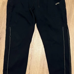 Svarta mjukisbyxor från Boss - Snygga svarta mjukisbyxor från Boss med diskret logga på sidan. Byxorna har en bekväm passform med snörning i midjan och stilrena sömmar längs benen. Perfekta för en avslappnad stil.