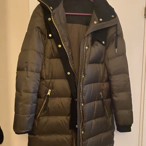 Brun/khaki dunjacka från J.Lindeberg - Snygg brun/khaki dunjacka från J.Lindeberg med huva och dragkedja. Jackan har långa ärmar och praktiska fickor med dragkedjor. Perfekt för kyliga dagar.